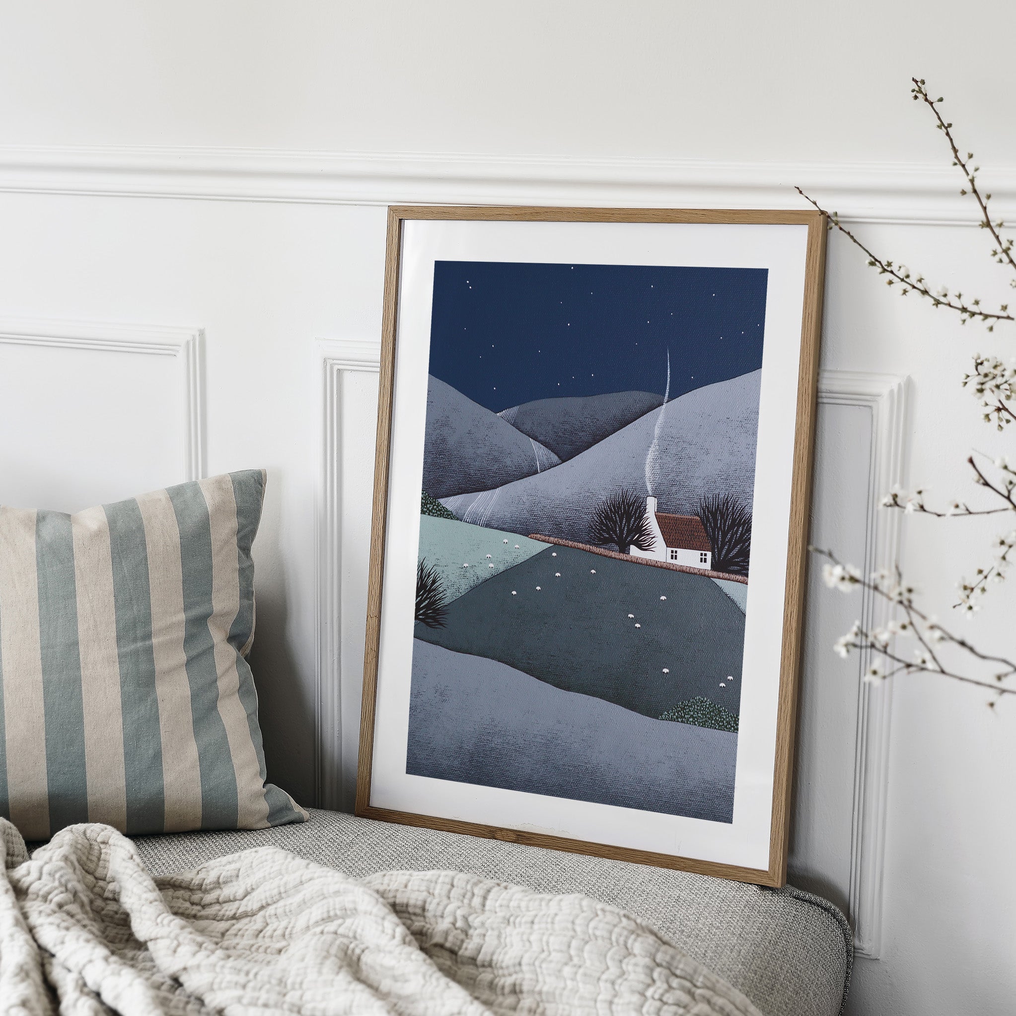 Art print featuring a cosy home nestled in rolling midnight hills in the evening countryside. Art print is leaning on a sofa in a living room.