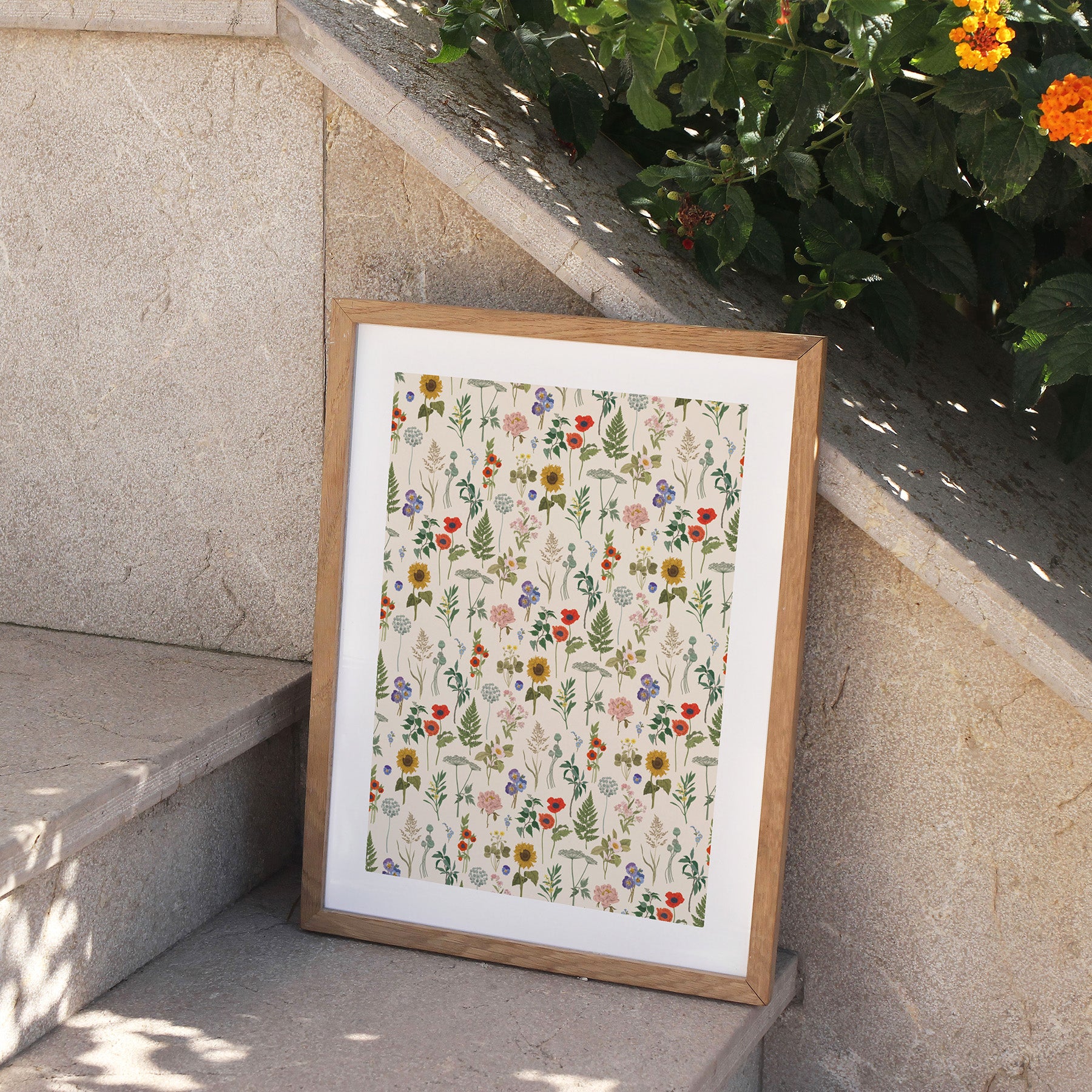 Framed floral pattern print on a stone ledge with plants in the background.