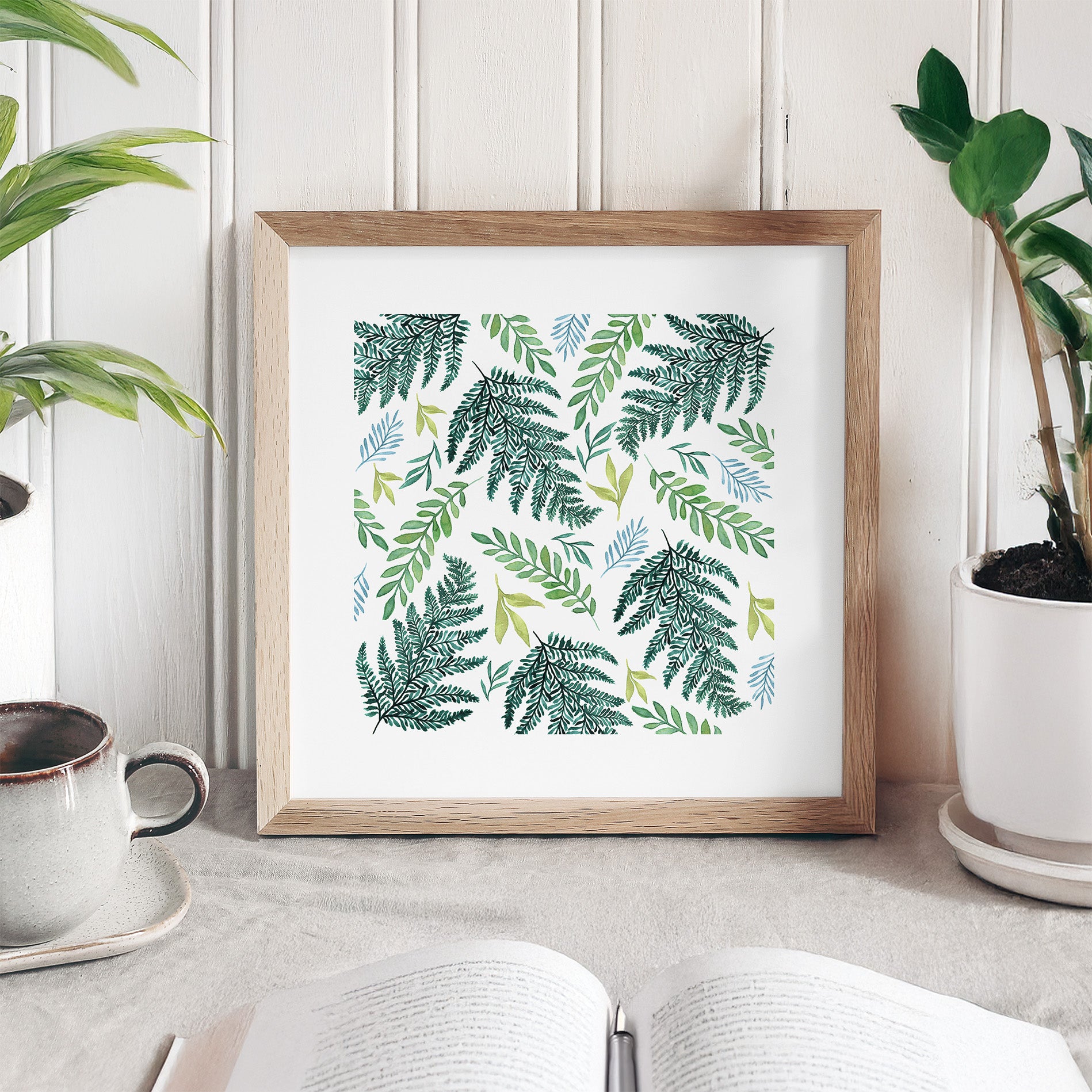Framed artwork with green leaf pattern on a table with plants and a cup.