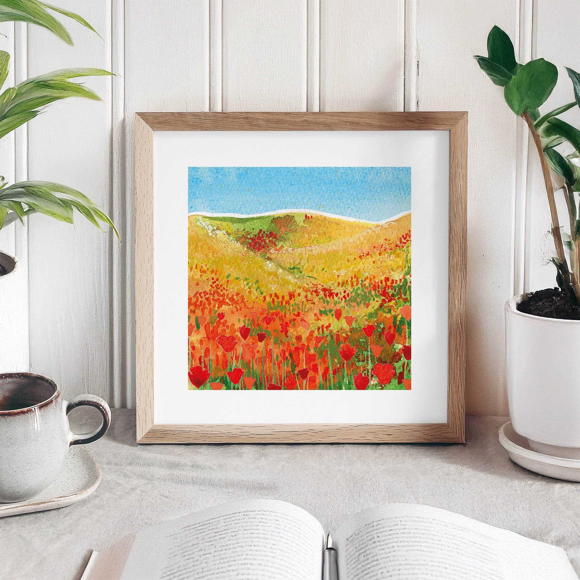 Framed artwork of a colorful landscape with red flowers on a white surface.