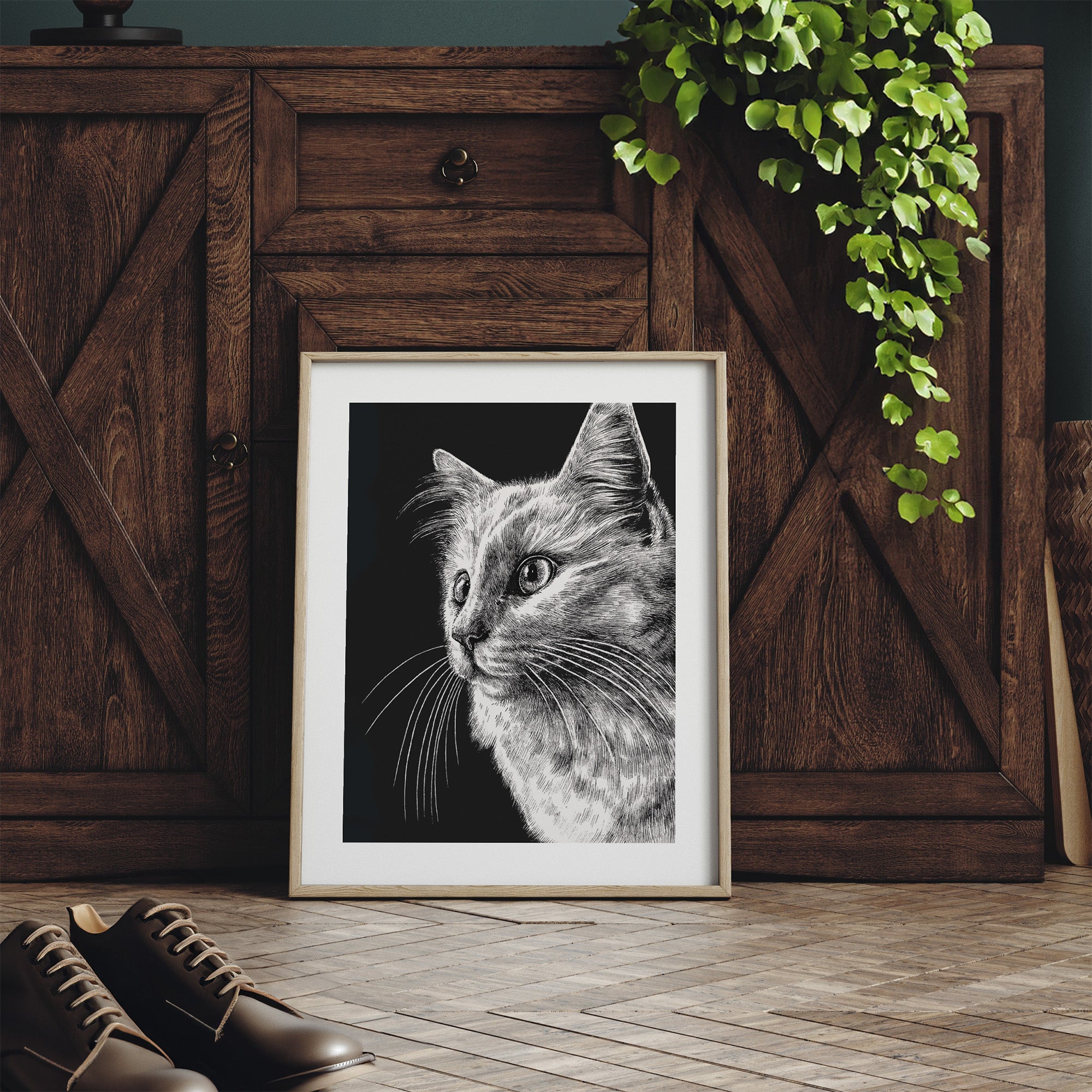 Framed black and white cat portrait on a wooden floor with a wooden cabinet and plant in the background.