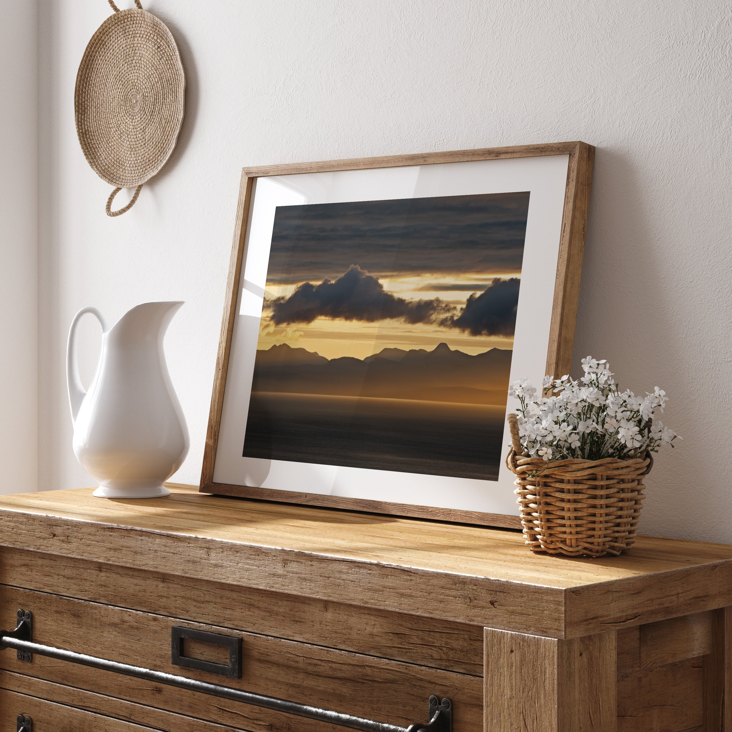 Framed photography art print featuring a sunset behind mountains near a lake. Print is leaning on a dresser against a white wall.