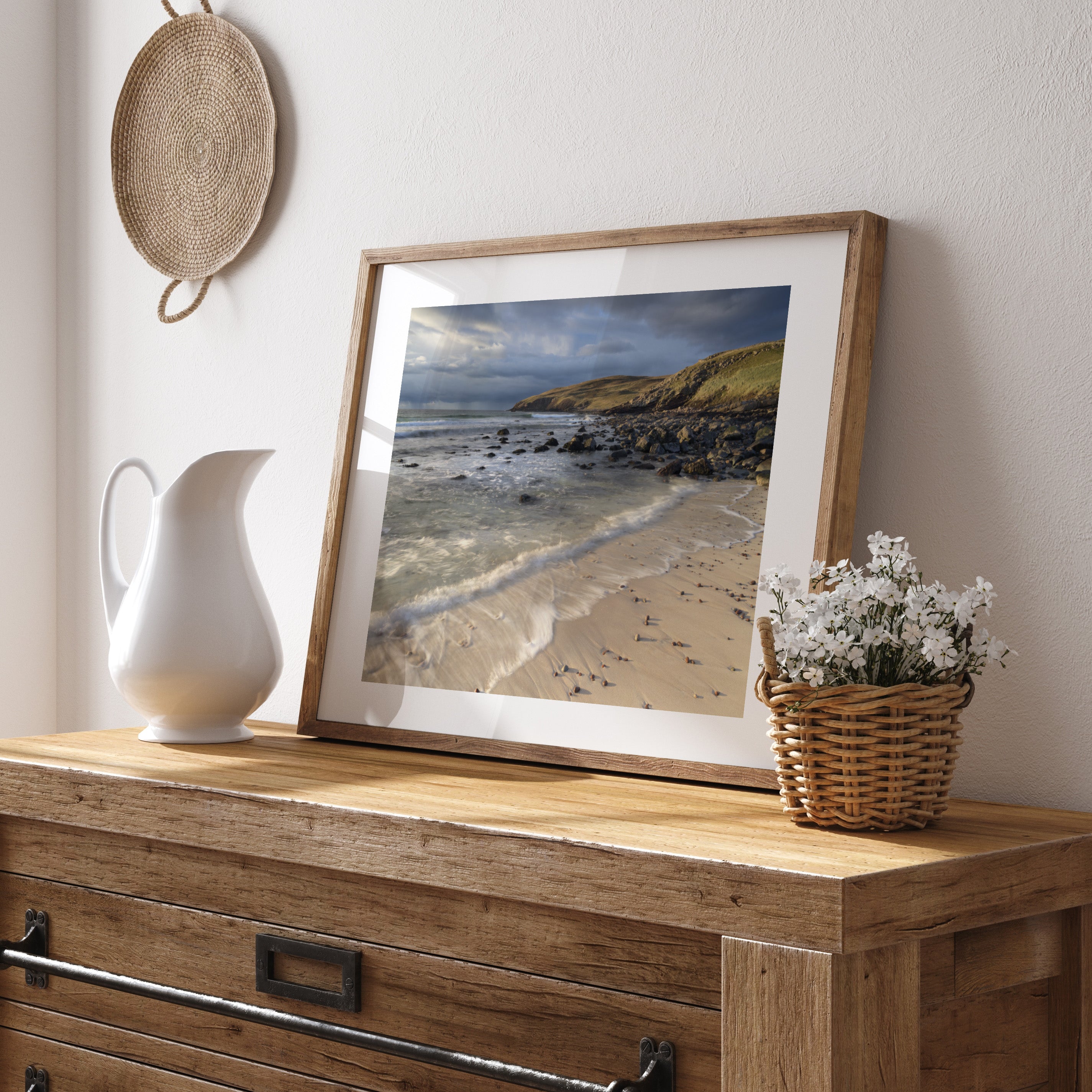 Framed photography art print of a beach and shoreline on a wooden dresser with a pitcher and basket.