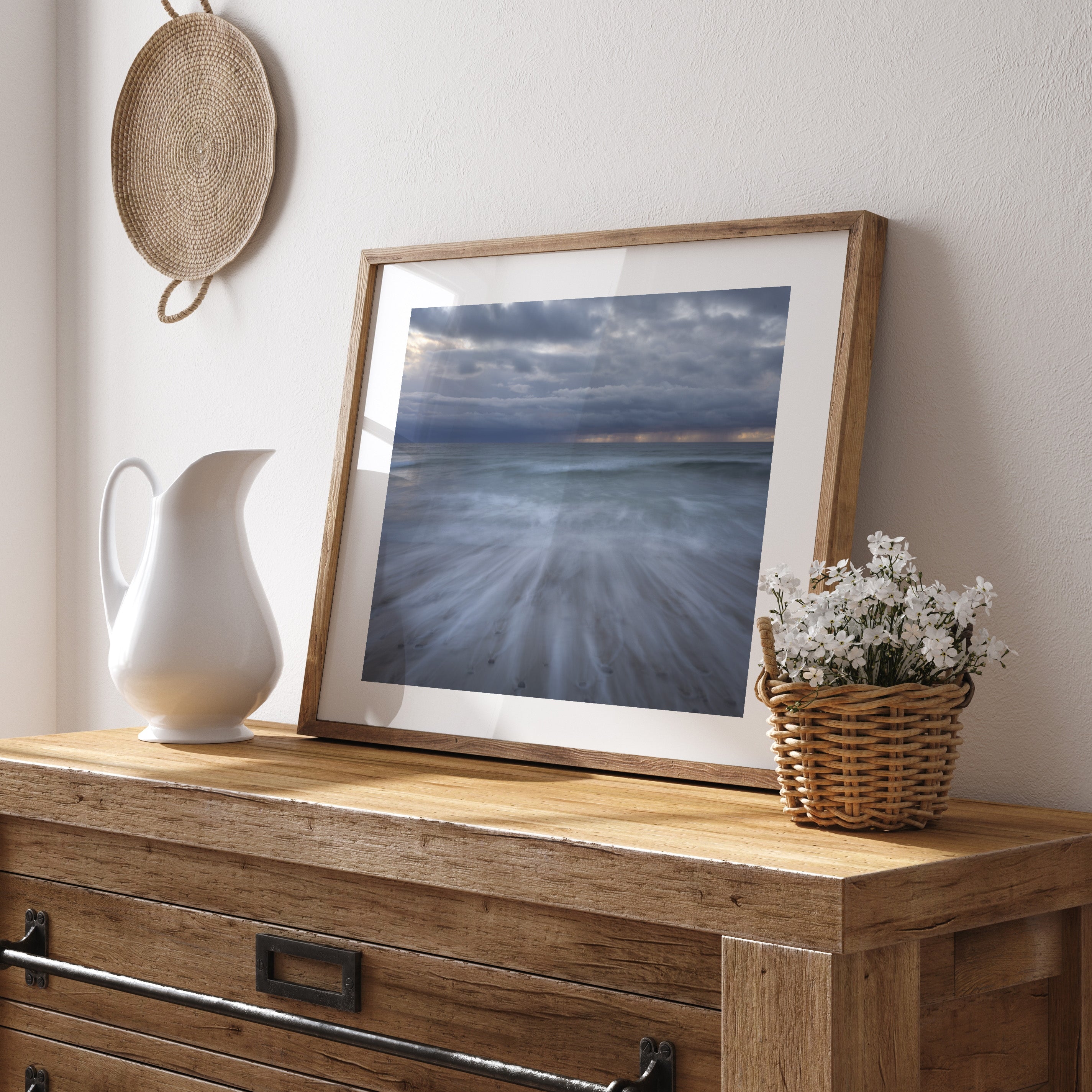 Wooden dresser with framed photography art print of a wave breaking on the shore, with a white pitcher, and basket of flowers against a light wall.