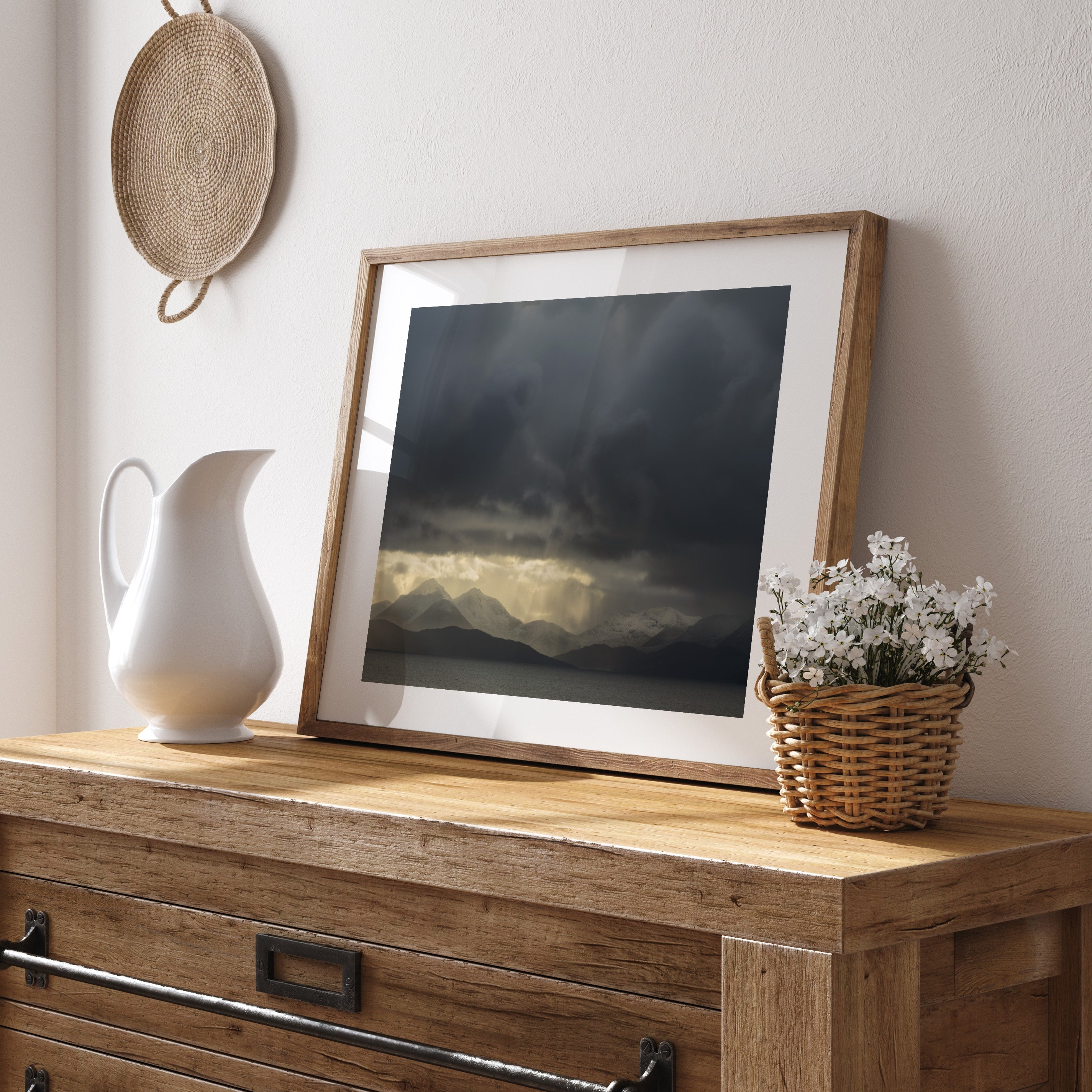 Framed photography art print featuring a storm breaking over wintery mountains, on a wooden dresser with a pitcher and basket.