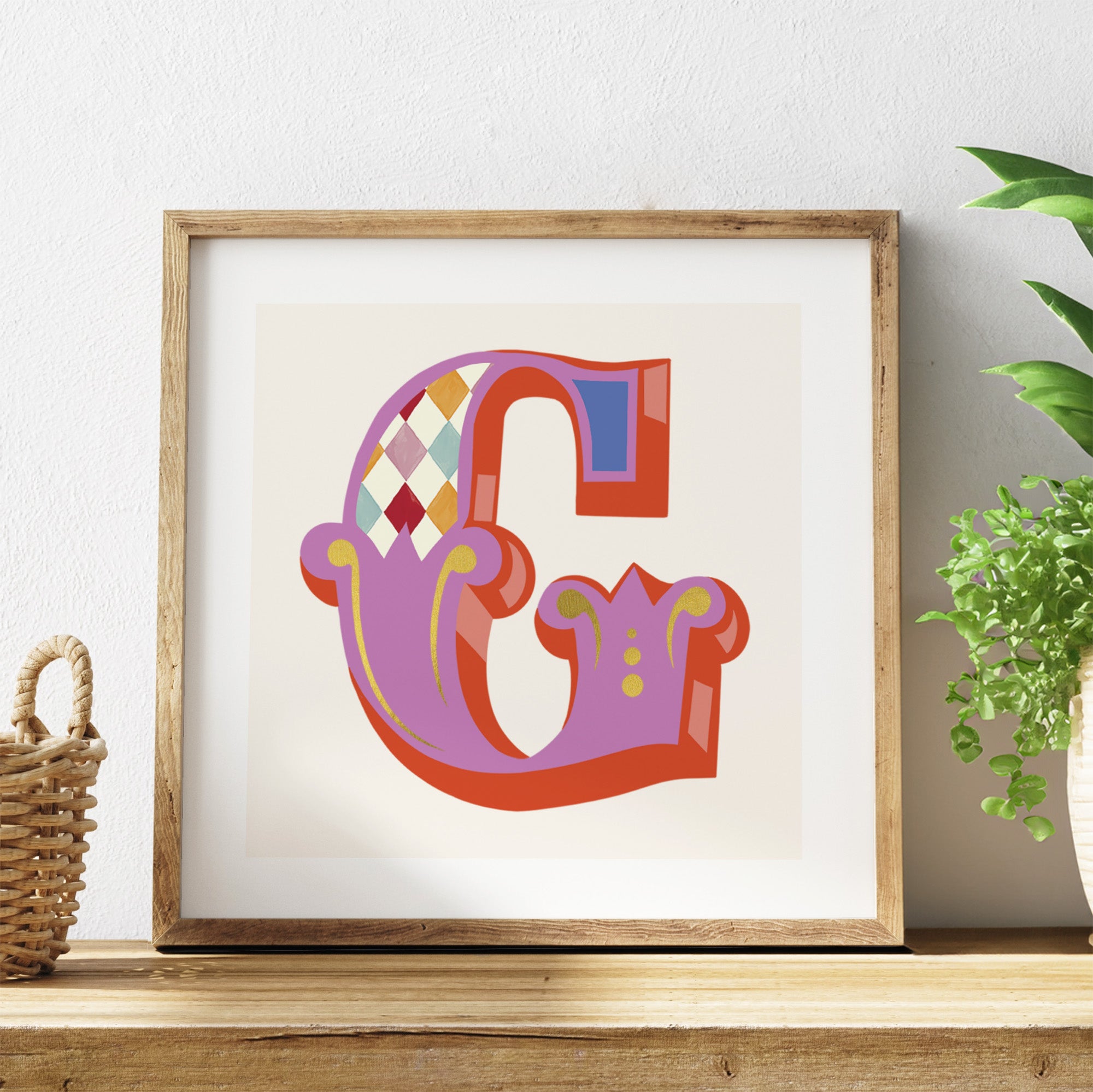Framed colourful letter 'G' on a shelf with a plant in the background.