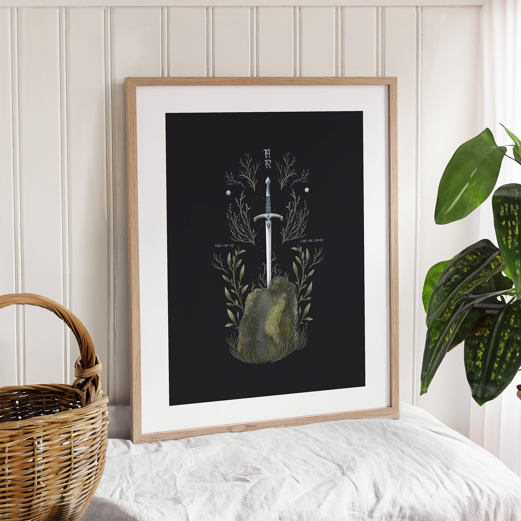A framed art print featuring the sword Excalibur coming out of the stone surrounded by botanicals on a black background. Print is leaning against a wall.