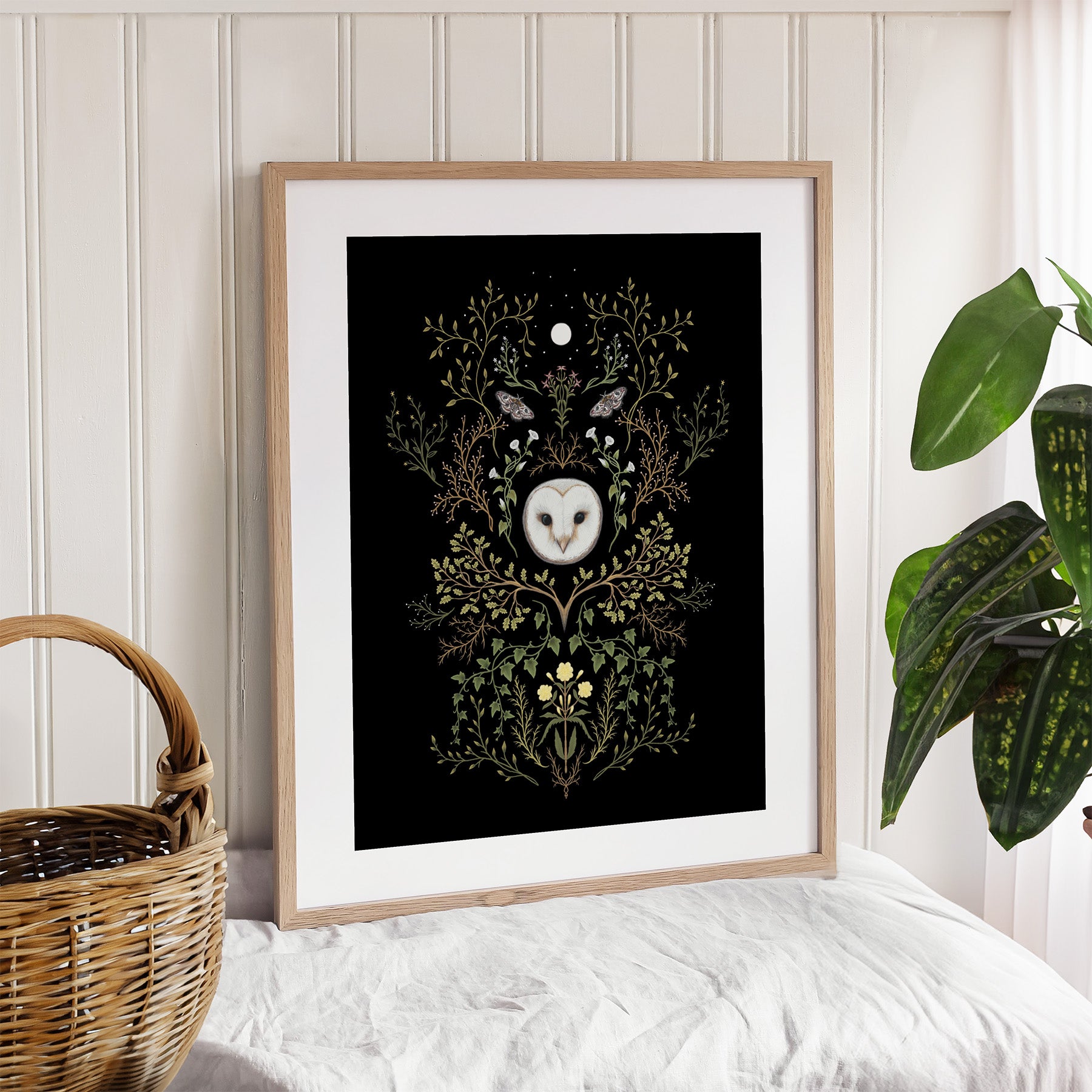 Framed artwork with an owl and moon design on a wall with a plant and basket.