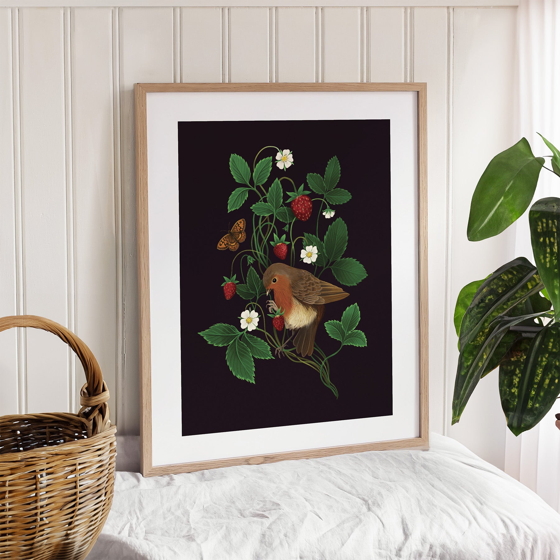 Framed art print featuring a robin perched within a strawberry plant on a black background. Print is leaning against a wall.