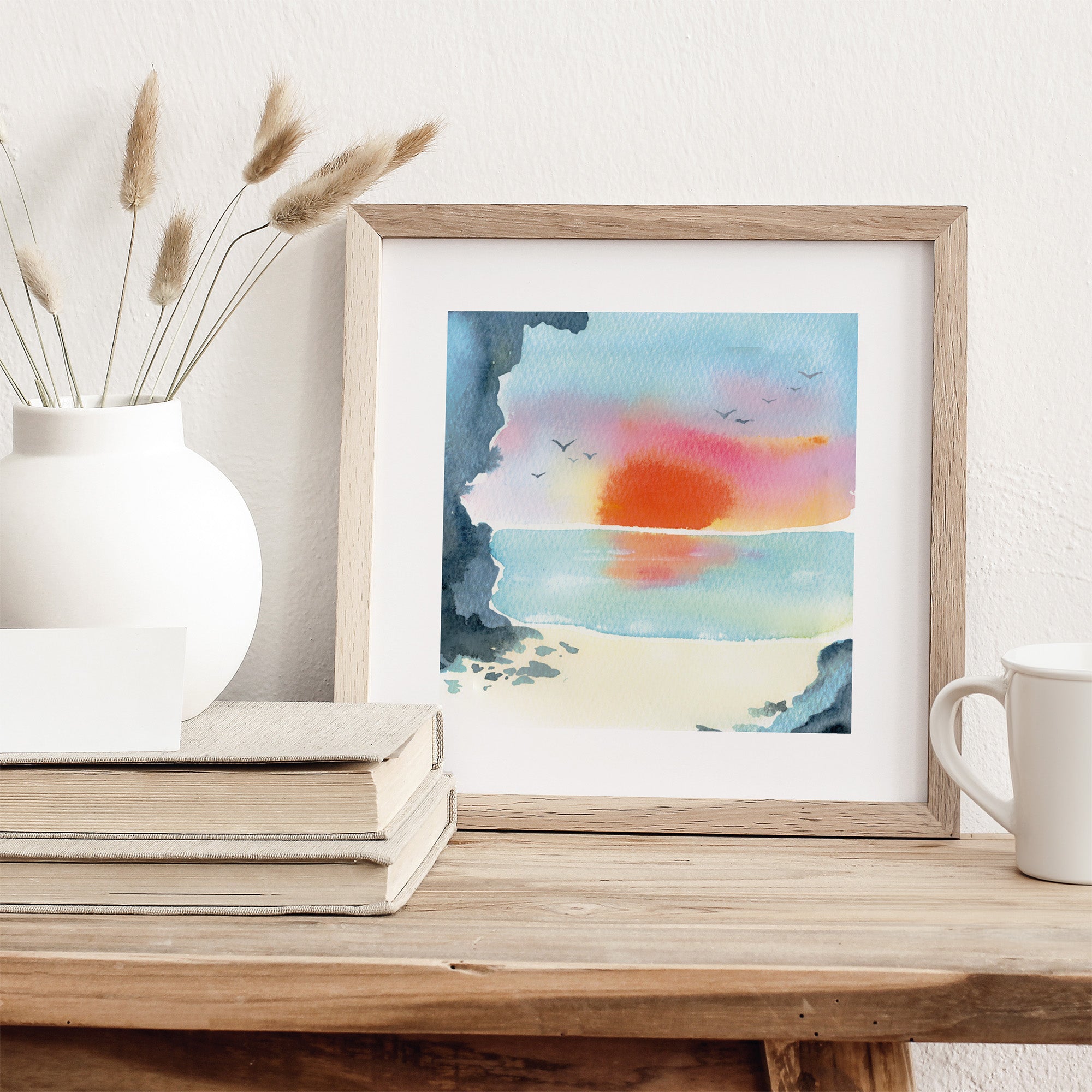 Framed watercolor painting of a sunset over a beach on a wooden table with a vase and books.